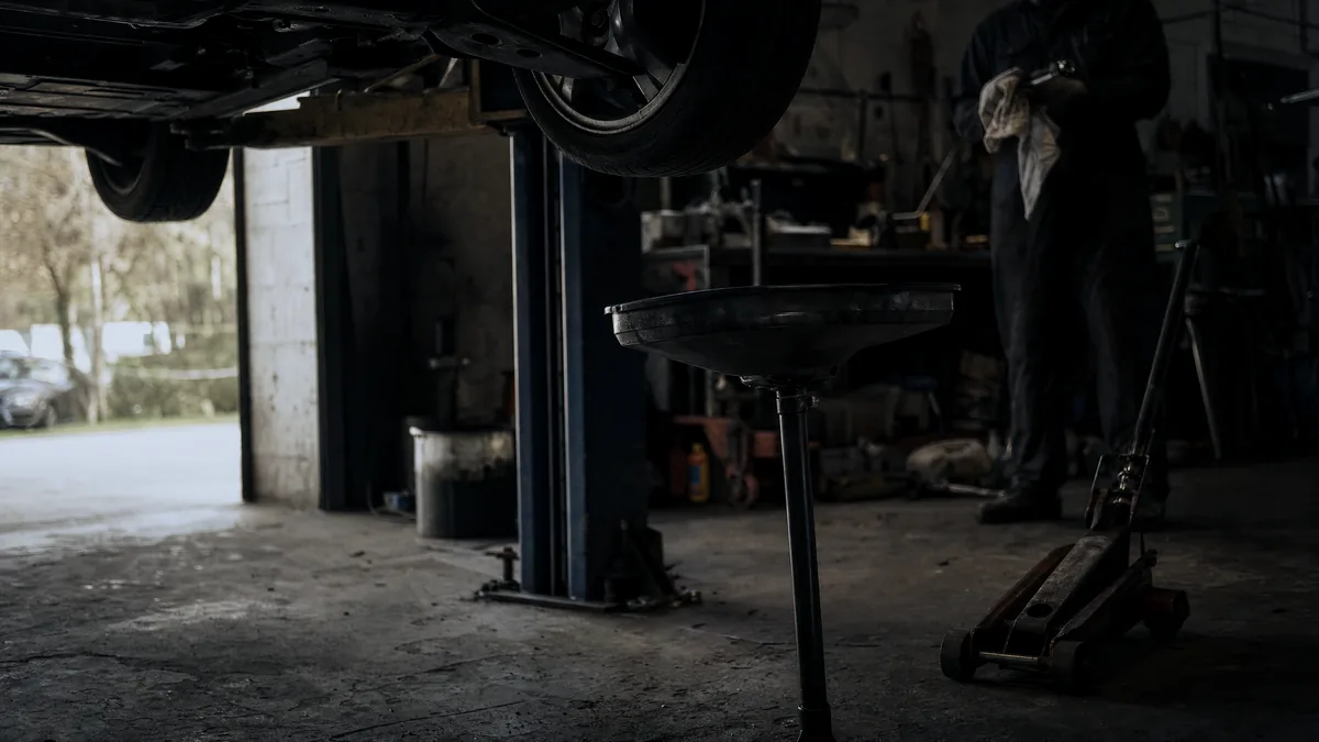 A car raised on a two-post lift in a dim garage, with an oil drainer and trolley jack on stained concrete and a mechanic wip
