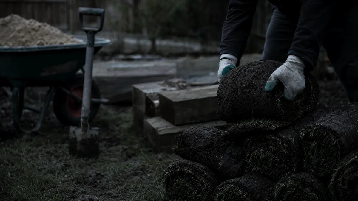 Gloved hands lift a roll of turf from a stack in a muddy garden, beside timber sleepers, a spade and a wheelbarrow