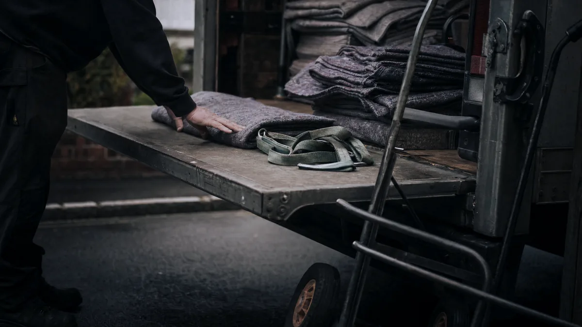 A worker's hand smooths a folded grey moving blanket on a removals van tailgate beside a sack truck