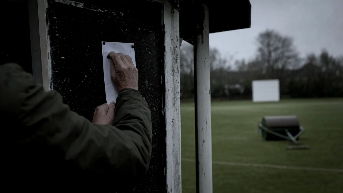 A hand pins a blank paper sheet to a weathered wooden pavilion board beside a damp cricket field and roller